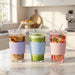 Three clear glass tumblers with ribbed silicone sleeves in lavender, light blue, and pink, each with a clear lid and straw, filled with iced coffee, green drink, and fruit punch on a marble kitchen counter.