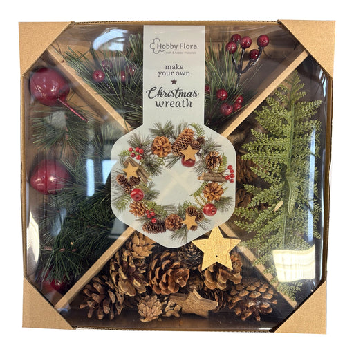 A Hobby Flora 'make your own Christmas wreath' kit in a clear-sided hexagonal box with a kraft cardboard base. The kit contains green pine branches, pinecones, red berries, a white wreath template with a printed wreath design, and gold star accents.